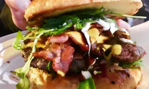 Soczysty burger z bekonem, serem, rukolą i sosami, trzymany w dłoni.
