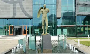 Brązowe rzeźby przedstawiające postacie ludzkie, w tym anioła, stoją przed nowoczesnym budynkiem z przeszkloną fasadą.