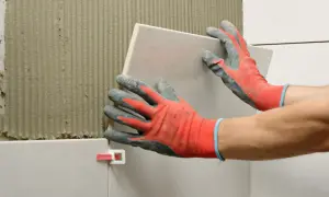 Układanie płytek 60x60. Ręce w rękawicach montują kolejną płytkę na ścianie z klejem. Sprawdź cennik układania płytek.