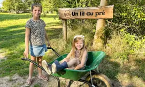 Deux petites filles profitent d'un séjour à la ferme, l'une assise dans une brouette, l'autre à côté, sous un panneau "Un lit au pré".