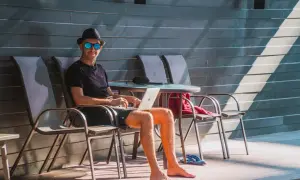 Mężczyzna w kapeluszu i okularach przeciwsłonecznych pracuje na laptopie na tarasie. Wygląda, jakby cieszył się dodatkowymi dniami urlopu.