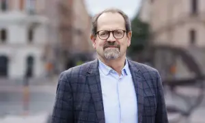 Peder Hasslev, en man med glasögon och skägg, bär en rutig kavaj och ljusblå skjorta. Bakgrunden är en suddig stadsmiljö.