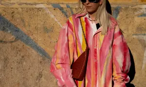 Kobieta w okularach przeciwsłonecznych i koszuli w stylu tie-dye w odcieniach różu i żółci, z brązową torebką na łańcuszku.