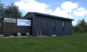Mobilny garaż bez pozwolenia, z banerem "Zbuduj sam dom", stoi na trawie.