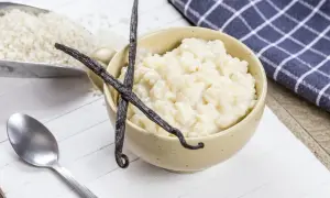Un bol de riz au lait crémeux, garni de gousses de vanille, avec du riz brut dans une pelle à côté.