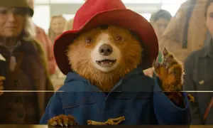 Pluszowy miś Paddington w czerwonym kapeluszu i niebieskim płaszczu, machający łapką.