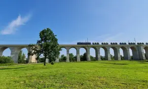 Pociąg przemierza najdłuższy most kolejowy w Polsce, majestatyczną kamienną konstrukcję z licznymi łukami, na tle błękitnego nieba i zielonej łąki.