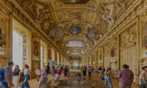 Prunkvolle Deckenmalereien und goldene Verzierungen im Louvre von innen. Besucher bestaunen die Kunstwerke in der langen Galerie.