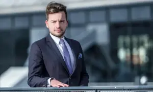Mężczyzna w garniturze i krawacie opiera się o barierkę na tle nowoczesnego budynku.