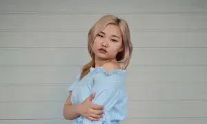 Młoda kobieta z blond włosami w niebieskiej sukience w paski, na tle białej ściany.