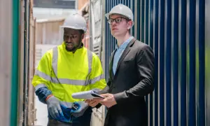 Due operai con caschi bianchi discutono documenti tra container. Uno indossa giacca alta visibilità, l'altro un completo. Verificano i permessi par.