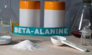 Biały proszek beta-alaniny na blacie, obok miarki i słoiczka. W tle dwa białe pojemniki z pomarańczowymi nakrętkami. Czy beta-alanina skutki uboczne są powodem do obaw?