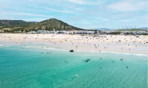Playa de Zahara de los Atunes con gente bañándose y disfrutando del sol, con el pueblo blanco al fondo y molinos de viento en la colina.