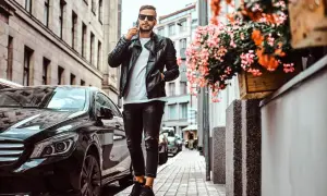 Mężczyzna w czarnej skórzanej kurtce i zniszczonych jeansach, rozmawiający przez telefon, obok lśniącego czarnego samochodu. Idealne buty do kurtki skórzanej dopełniają stylizację.
