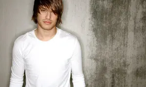 Michael Tyler, a young man with tousled brown hair and a white long-sleeved shirt, poses against a textured concrete wall.