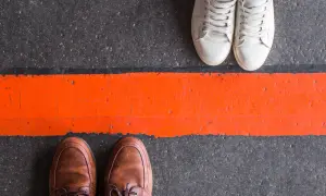 Dwie pary butów, jedna w brązowych, druga w białych, stoją po przeciwnych stronach pomarańczowej linii. To metafora tego, jak stawiać granice.