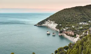 Baia con scogliere bianche e mare azzurro, perfetta per una passeggiata dell'amore. Alberi verdi e case bianche punteggiano la costa.