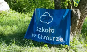 Niebieski baner z napisem "Szkoła w Chmurze" oparty o drzewo, na tle zielonej trawy i liści.