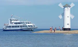 Biała latarnia morska z wiatrakami na tle błękitnego nieba. Obok niej cumuje statek wycieczkowy, a na brzegu odpoczywają ludzie.