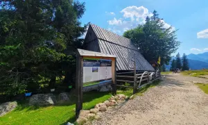 Drewniana chata na górskim szlaku, obok tablica informacyjna o kulturowym wypasie owiec. W oddali widać wierch Poroniec.