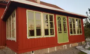 Ett rött hus med pågående arbete för att bygga glasveranda. Gröna fönster och dörrar reflekterar himlen.