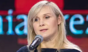 Kobieta z blond włosami mówi do mikrofonu na tle ekranu z napisem.
