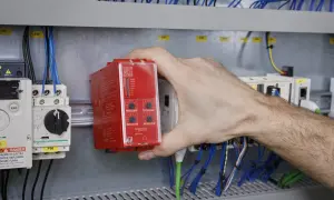 Ręka instalatora montuje czerwony sterownik bezpieczeństwa w szafie sterowniczej, wśród przewodów i innych modułów.