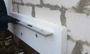 Ręka w rękawiczce sprawdza poziomowanie styropianu przy ścianie z bloczków. Kołkowanie styropianu trwa.