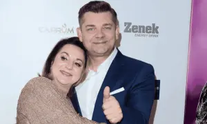 Danuta Martyniuk, w złotej sukience, przytula się do mężczyzny w granatowym garniturze, który pokazuje kciuk w górę.