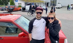 Ett par poserar framför en röd bil, redo att bygga ljud i bil. I bakgrunden syns bilar och folk vid en butik.
