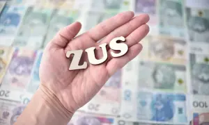 Drewniane litery ZUS leżą na dłoni, na tle polskich banknotów.