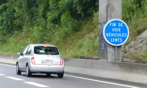 Voiture argentée sur l'autoroute, passant devant un panneau "Fin de voie véhicules lents". La nature luxuriante borde la route.