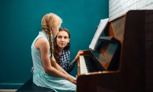 Dziewczynka z warkoczem uczy się gry na pianinie pod okiem nauczycielki. Nuty na pulpicie wskazują na lekcję muzyki.