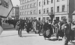 Människor med armbindlar ledsas förbi Schindlers fabrik. En soldat går framför.