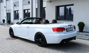 Białe BMW E93 cabrio z brązową skórzaną tapicerką i czarnymi felgami stoi na kostce brukowej przed nowoczesnym budynkiem.