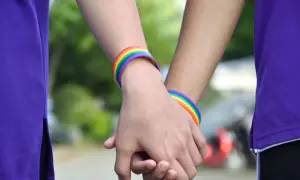 Zwei Hände halten sich, geschmückt mit Regenbogenarmbändern. Eine Person trägt ein lila Oberteil. Ein Symbol für Liebe, das halb Mann halb Frau sein kann.