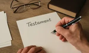 Ręka pisze piórem na kartce "Testament". Obok leżą okulary i książka. Dowiedz się, jak napisać testament bez notariusza.