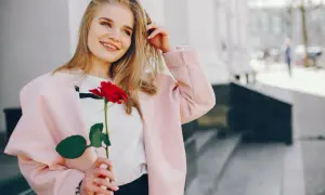 Uśmiechnięta blondynka w różowym płaszczu trzyma czerwoną różę. Idealna stylizacja, jak się ubrać na randkę.