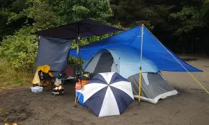 Campingplats med tält, eldstad och paraply. Ved torkar under presenning.