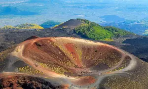 Widok na krater wulkanu Etna, gdzie ścieżka prowadzi turystów na wejście na etnę. W oddali zielone wzgórza i miasto.