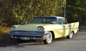 Gul Chrysler Saratoga från 1950-talet, med kromdetaljer och vita däcksidor, parkerad vid en lummig buske.