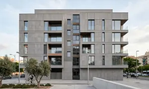 Edificio moderno con revestimiento fachada ventilada de piedra gris, balcones acristalados y detalles en madera. Un ciclista pasa frente a los olivos.