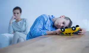 Chłopiec z zespołem Aspergera bawi się zabawką, podczas gdy matka w tle wygląda na zmartwioną, być może zmagając się z jego agresją.