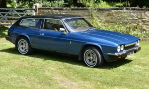 Blå Reliant Scimitar GTE, en klassisk sportsvagn med kombikaross, parkerad på en gräsmatta framför en stenmur och en grind.