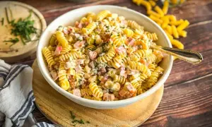 Schüssel mit cremigem Nudelsalat, gefüllt mit Schinken, Erbsen und Kräutern. Ein Löffel liegt darin. Dieser ist der beste Nudelsalat!