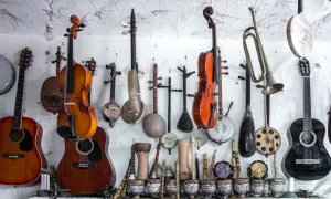 Różnorodne instrumenty strunowe, w tym gitary, skrzypce i tradycyjne lutnie, wiszą na ścianie obok bębnów i fletów.