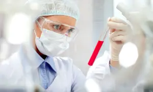 Naukowczyni w okularach i masce analizuje czerwoną ciecz w probówce. Badania pct.
