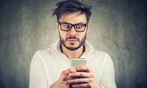 Przerażony mężczyzna w okularach czyta na telefonie o tym, co grozi za oczernianie drugiej osoby.
