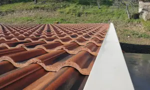 Tegole per tetti a bassa pendenza, color terracotta, disposte in file ordinate su un tetto. Sullo sfondo, un pendio erboso.