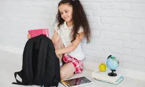 Dziewczynka pakuje różowy zeszyt do czarnego plecaka. Obok leżą tablet, jabłko i globus. Zastanawia się, jaki plecak najlepiej kupić do szkoły.